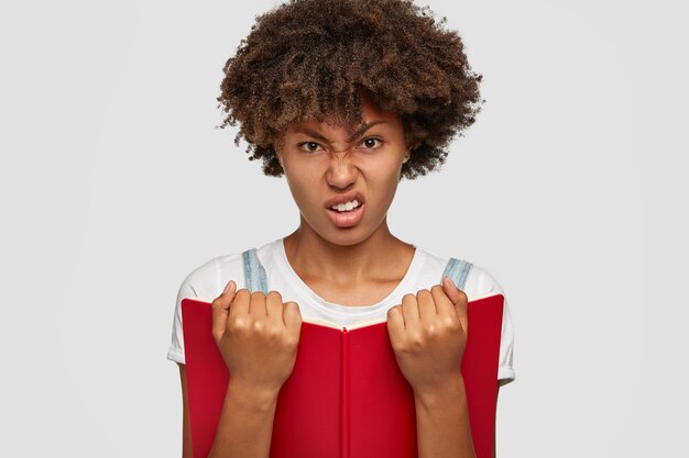 La foto di uno studente universitario insoddisfatto non ha alcun desiderio e non desidera studiare, tiene in mano il libro rosso, aggrotta le sopracciglia, si sente male e stanco di leggere, infastidito, modella su sfondo bianco. Persone, concetto di apprendimento