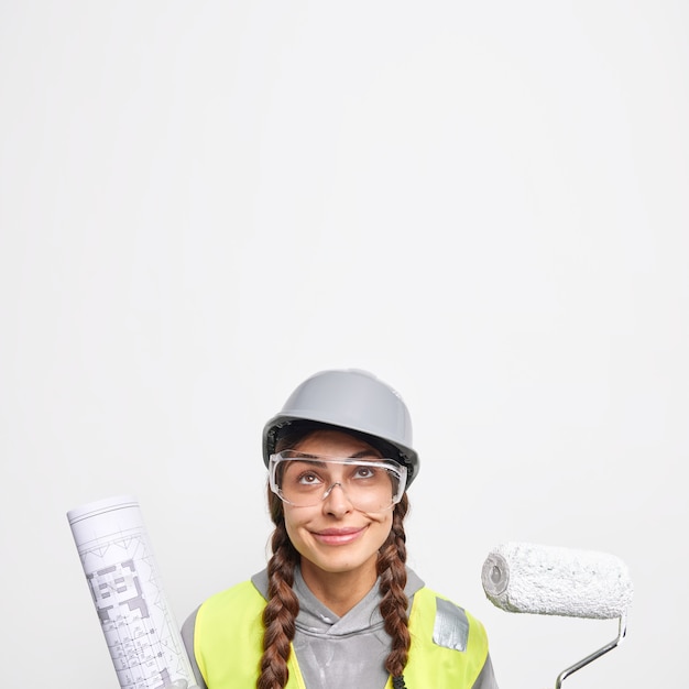 La foto in interni di una donna ingegnere soddisfatta tiene il rullo di verniciatura e il progetto di carta guarda sopra i lavori in cantiere
