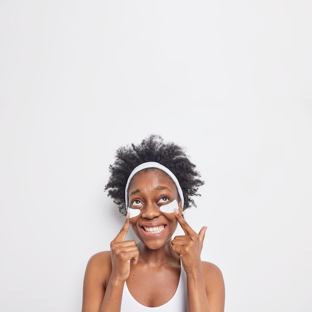 La foto verticale di una donna afroamericana riccia felice punta a sotto gli occhi sorrisi ampiamente