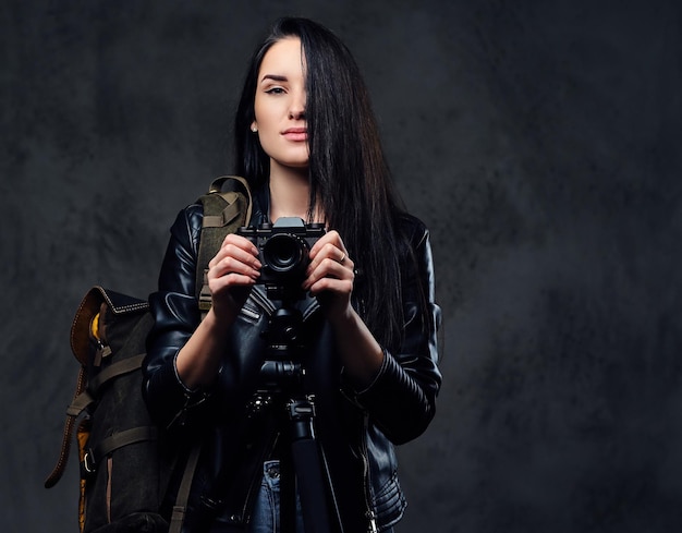La fotografa bruna tiene una fotocamera professionale su un treppiede e uno zaino da viaggio.