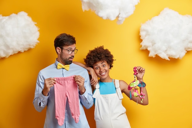 La futura mamma e il padre posano con giocattoli e vestiti per il bambino, preparati a diventare presto genitori. Futura mamma nel suo tardo periodo di gravidanza, tiene il cellulare, ha una pancia grande, isolata sul muro giallo.