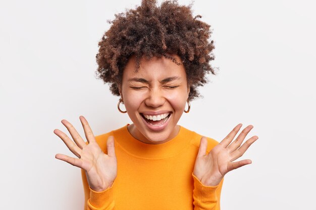 La gioiosa donna afroamericana esprime i sorrisi di felicità tiene ampiamente gli occhi chiusi alza i palmi ha un umore ottimista invia vibrazioni positive vestite casualmente isolate sul muro bianco. Concetto di emozioni