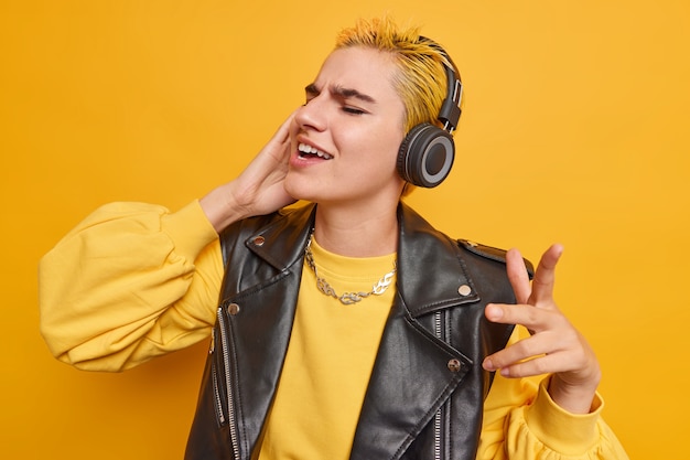 La gioiosa ragazza caucasica con l'acconciatura alla moda ascolta musica in cuffie wireless cattura ogni pezzo di canzone balla a ritmo vestita con abiti eleganti isolati sul muro giallo