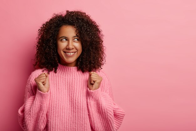 La giovane adolescente afroamericana di successo celebra la realizzazione, alza i pugni serrati, vestita con un maglione invernale oversize, sorride ampiamente, guarda da parte, isolato su sfondo rosa.