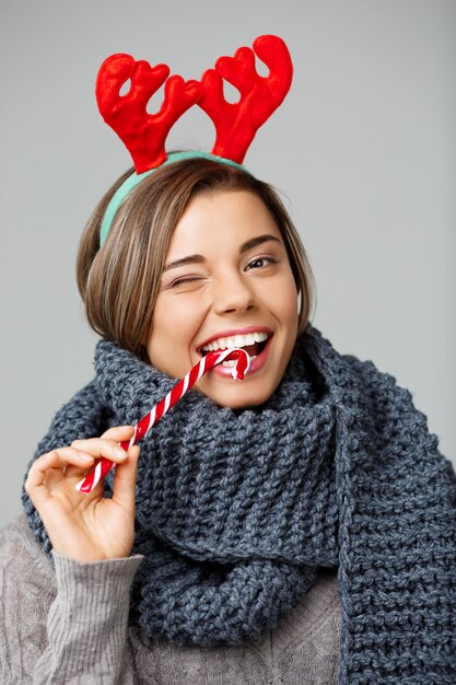 La giovane bella donna bionda in grandi corna tricottate della renna di natale e della sciarpa che sorride mangiando il lollypop a strisce su gray.