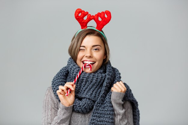 La giovane bella donna bionda in grandi corna tricottate della renna di natale e della sciarpa che sorride mangiando il lollypop a strisce su gray.