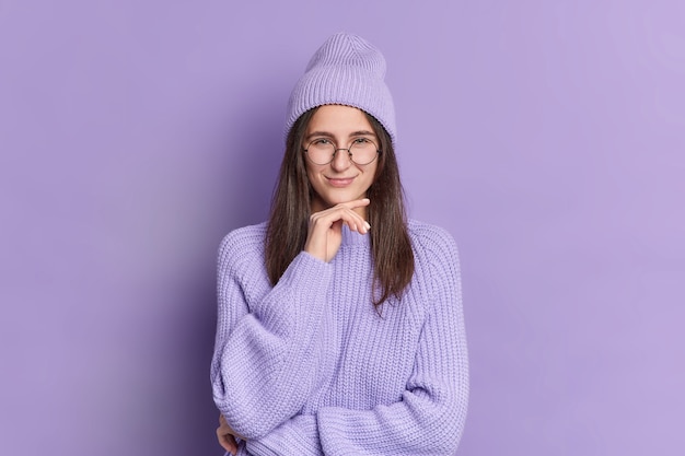 La giovane bella donna castana tiene il mento e ha un piano ingannevole di espressione sorniona sorride piacevolmente indossa un maglione lavorato a maglia cappello alla moda