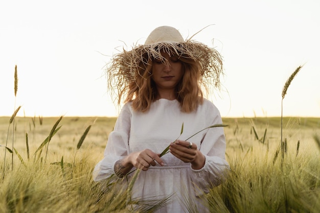 la giovane bella donna con i capelli lunghi biondi in un vestito bianco in un cappello di paglia raccoglie i fiori su un campo di grano. Capelli volanti al sole, estate. Tempo per sognatori, tramonto dorato.