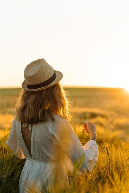 la giovane bella donna con i capelli lunghi biondi in un vestito bianco in un cappello di paglia raccoglie i fiori su un campo di grano. Capelli volanti al sole, estate. Tempo per sognatori, tramonto dorato.