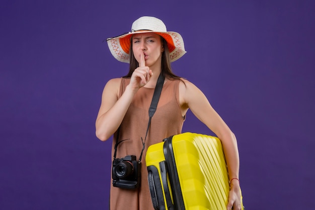 La giovane bella donna del viaggiatore in cappello dell'estate con la valigia gialla e la macchina fotografica che fanno il silenzio gesticolano con il dito sulle labbra sopra la parete porpora