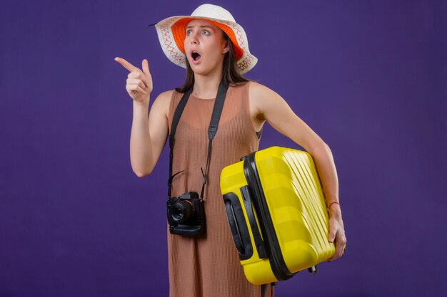 La giovane bella donna del viaggiatore in cappello dell'estate con la valigia gialla e la macchina fotografica che indicano qualcosa che guarda con l'espressione di paura ha stupito sopra la parete porpora