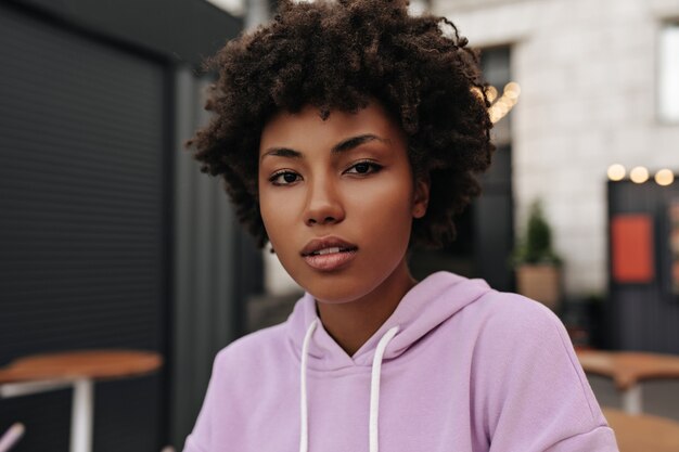 La giovane bella donna riccia castana in felpa con cappuccio viola esamina la macchina fotografica