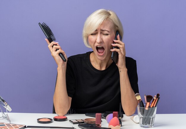 La giovane bella ragazza arrabbiata si siede alla tavola con gli strumenti di trucco parla sul pettine della tenuta del telefono isolato sulla parete blu