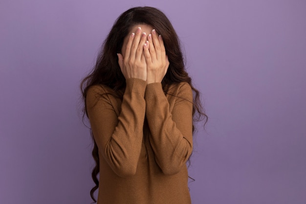 La giovane bella ragazza che indossa il maglione a collo alto marrone ha coperto il viso con le mani isolate sul muro viola