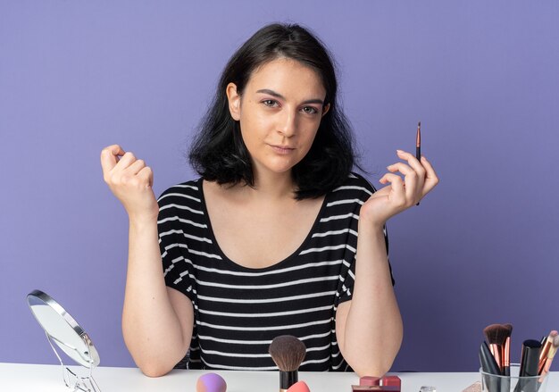 La giovane bella ragazza dall'aspetto impressionato si siede al tavolo con gli strumenti per il trucco che tengono il pennello per il trucco isolato sulla parete blu