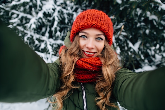 La giovane bella ragazza felice in cappello e sciarpa tricottati inverno rosso prende il selfie