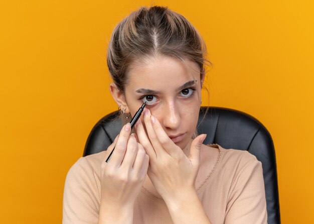 la giovane bella ragazza si siede al tavolo con gli strumenti per il trucco disegna la freccia con l'eyeliner isolato sul muro arancione