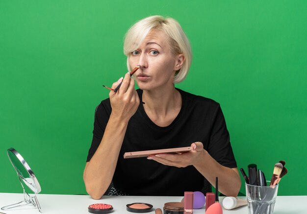 la giovane bella ragazza si siede alla tavola con gli strumenti di trucco che applicano l'ombretto con la spazzola di trucco isolata sulla parete verde