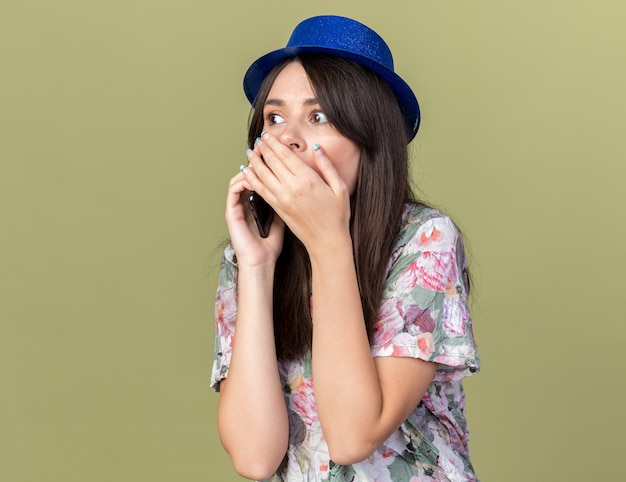 La giovane bella ragazza sorpresa che indossa il cappello del partito parla sul telefono ha coperto la bocca di mano isolata sulla parete verde oliva