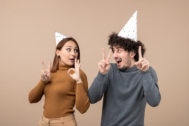 La giovane coppia adorabile indossa la ragazza sorridente del cappello del nuovo anno che mostra gli occhiali e il gesto di vittoria e il ragazzo eccitato divertente