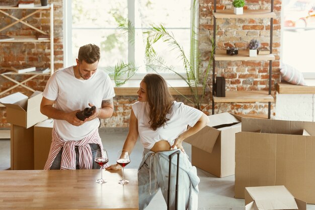 La giovane coppia si è trasferita in una nuova casa o appartamento. Bere vino rosso, sorridere e rilassarsi dopo aver pulito e disimballato. Sii felice e fiducioso. Famiglia, trasloco, relazioni, concetto di prima casa.