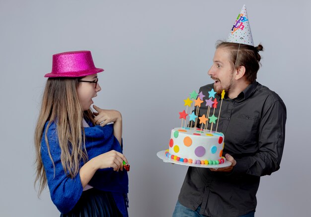 La giovane coppia sorpresa guarda a vicenda la ragazza con gli occhiali che porta il cappello rosa tiene il fischio e l'uomo bello nella torta della tenuta del cappuccio di compleanno isolata sulla parete bianca