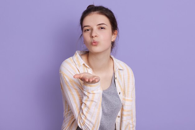 La giovane donna affascinante del brunette che porta l'abbigliamento casuale che mostra il bacio dell'aria, mantiene le labbra arrotondate