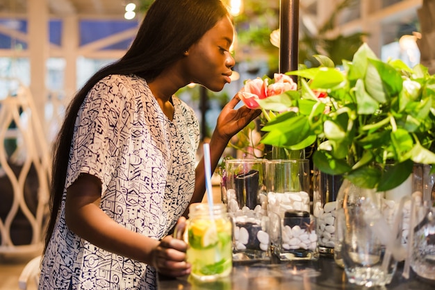 La giovane donna afroamericana allegra in vestito da estate al caffè annusa i fiori bianchi in vaso.