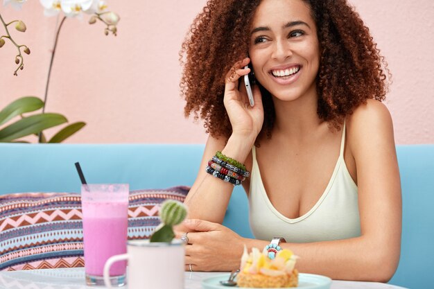 La giovane donna afroamericana dalla pelle scura positiva ha una conversazione piacevole tramite un moderno smart phone, ricrea nella caffetteria, beve frullato e mangia la torta