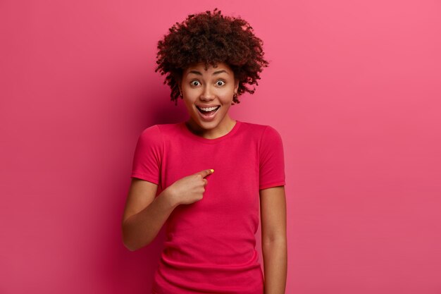 La giovane donna afroamericana positiva indica se stessa con eccitazione, ha una reazione felice e inaspettata, chiede se mi prendi in giro, ride positivamente, indossa una maglietta rossa, posa contro il muro rosa