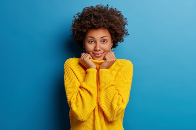 La giovane donna afroamericana positiva sorride ampiamente e indossa un maglione giallo isolato su sfondo blu.