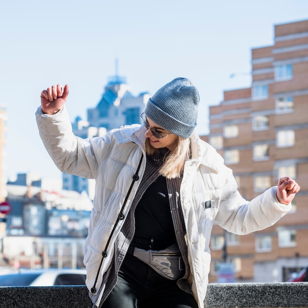 La giovane donna alla moda che indossa il blu tricotta il dancing del cappello nella città