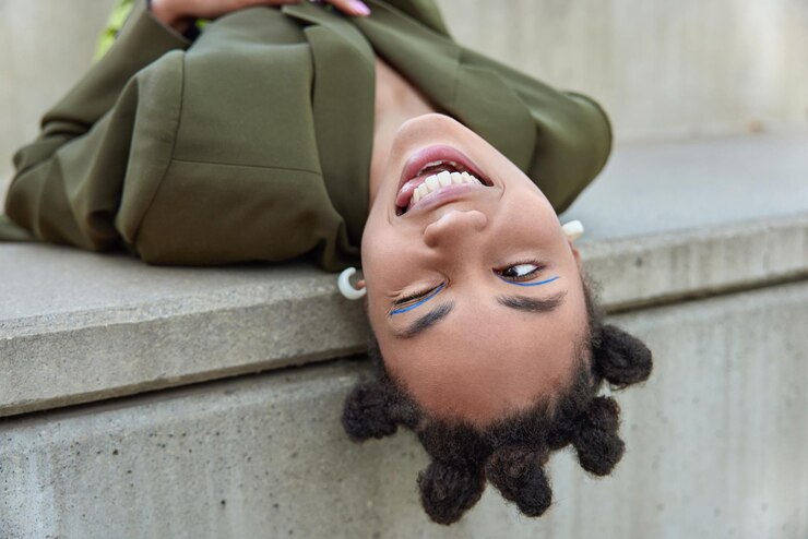 La giovane donna alla moda giocosa con i panini per capelli strizza l'occhio agli occhi sporge la lingua vestita con una giacca verde ha un trucco vivido che mostra i denti bianchi pieni di energia. Emozioni delle persone e concetto di stile