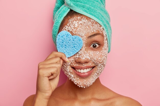 La giovane donna allegra copre un occhio con una spugna cosmetica a forma di cuore, rimuove il trucco, ha uno scrub al sale marino intorno al viso, tenero sorriso