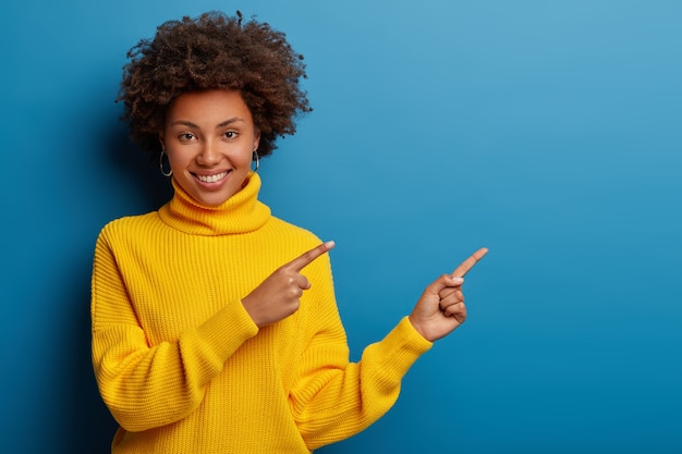 la giovane donna allegra in maglione giallo indica da parte nello spazio della copia, mostra la pubblicità fredda contro fondo blu