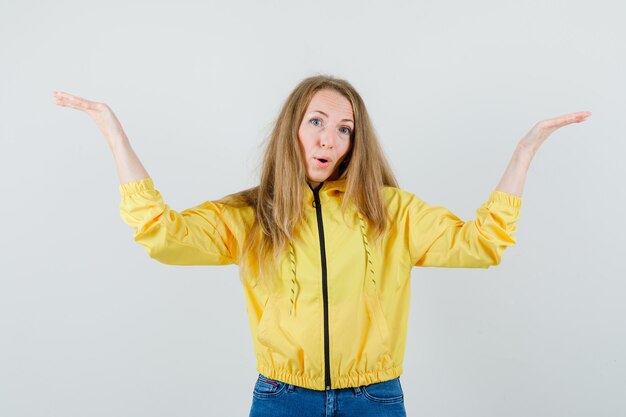 La giovane donna allunga le braccia come esultante in bomber giallo e jeans blu e sembra felice. vista frontale.