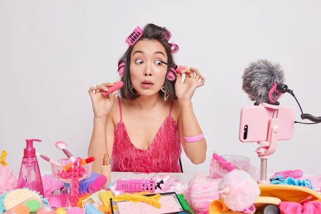 La giovane donna asiatica applica il mascara presenta un nuovo prodotto di bellezza registra il video in livestreaming si siede al tavolo disordinato indossa bigodini vestito rosa isolato su bianco