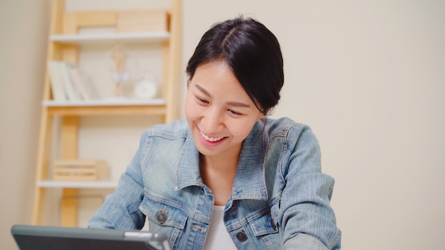 La giovane donna asiatica che lavora facendo uso della compressa che controlla i media sociali mentre si rilassa sullo scrittorio in salone a casa.