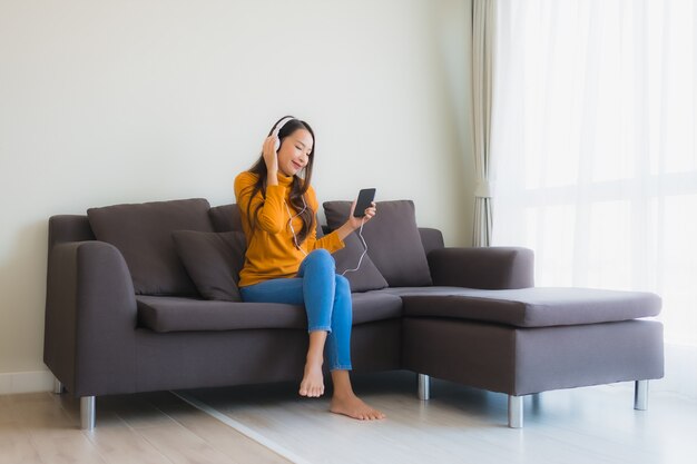 La giovane donna asiatica che per mezzo dello smartphone con le cuffie per ascolta musica sul sofà