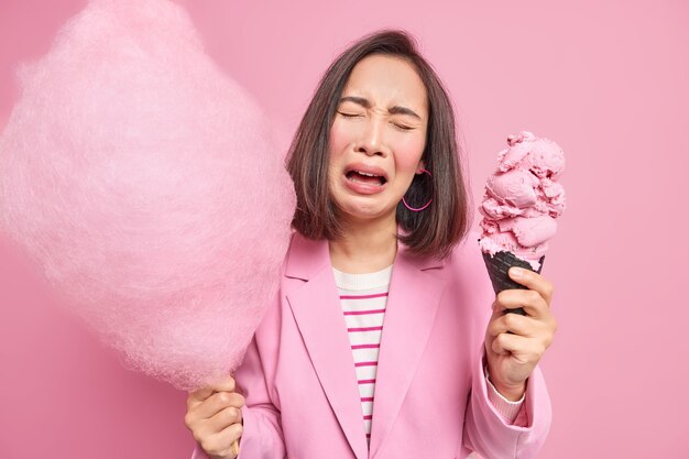 La giovane donna asiatica che piange sconsolata con i capelli scuri tiene in mano zucchero filato dolce e il gelato sente la tentazione di mangiare un dessert ad alto contenuto calorico che continua a seguire le pose della dieta al coperto. Concetto di dieta e cibo spazzatura ju