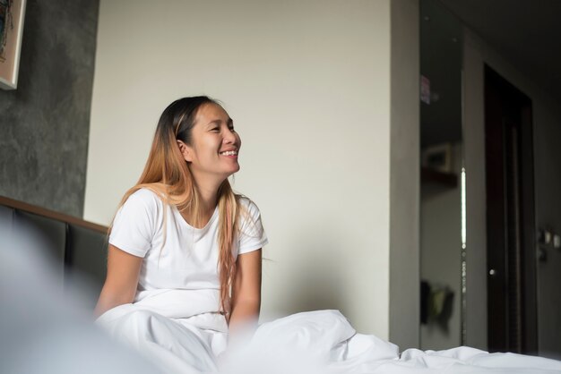 La giovane donna asiatica che respira e che si siede sorride su un letto