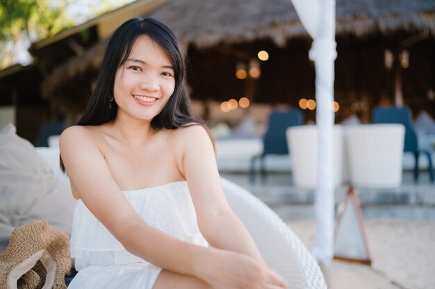 La giovane donna asiatica che si siede sul banco si rilassa sulla spiaggia, bella femmina felice si rilassa vicino al mare.