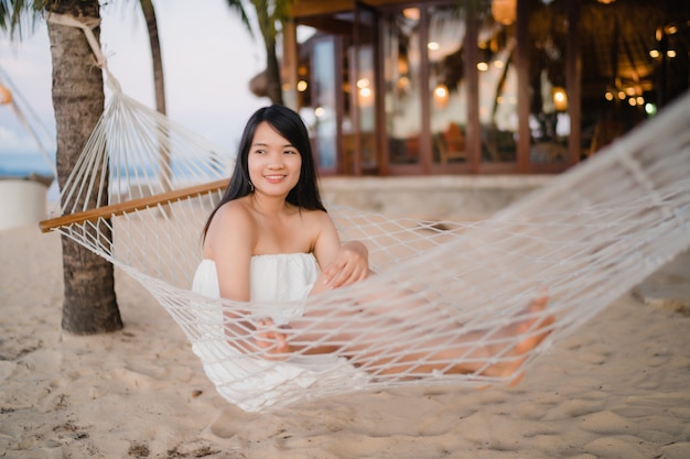 La giovane donna asiatica che si siede sull&#39;amaca si rilassa sulla spiaggia, bella femmina felice si rilassa vicino al mare.