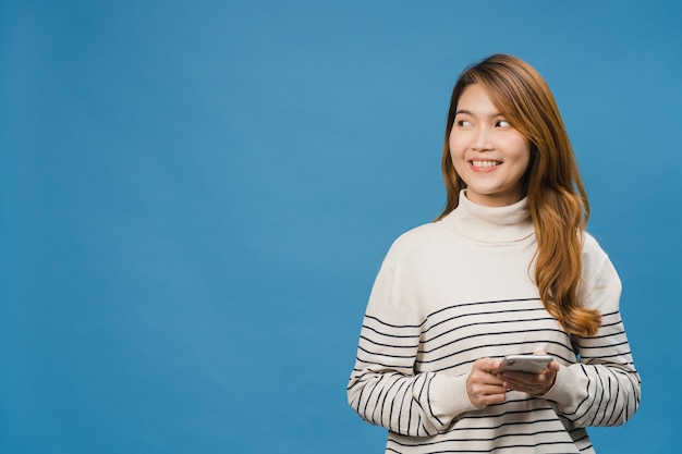 La giovane donna asiatica che usa il telefono con un'espressione positiva, sorride ampiamente, vestita con abiti casual sentendosi felice e in piedi isolata sul muro blu