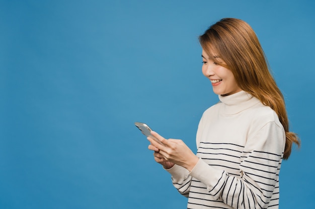 La giovane donna asiatica che usa il telefono con un'espressione positiva, sorride ampiamente, vestita con abiti casual sentendosi felice e in piedi isolata sul muro blu