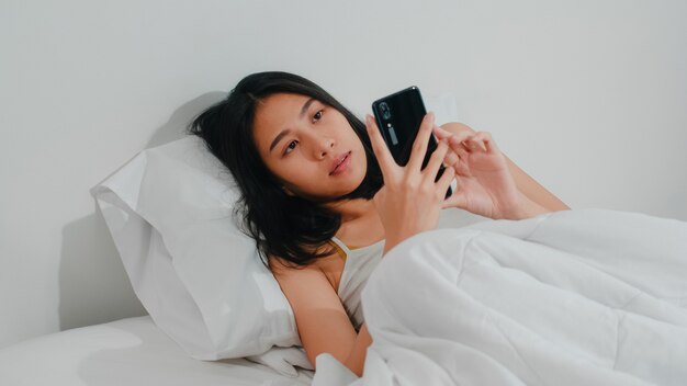 La giovane donna asiatica che utilizza lo smartphone che controlla i media sociali che ritiene sorridere felice mentre si trova sul letto dopo sveglia di mattina, la bella signora ispanica attraente che sorride si rilassa nella camera da letto a casa.