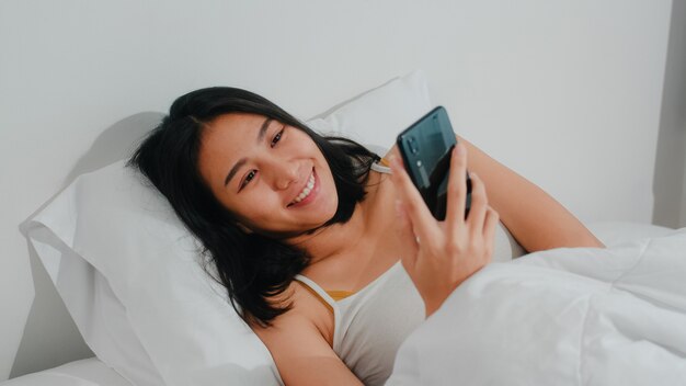 La giovane donna asiatica che utilizza lo smartphone che controlla i media sociali che ritiene sorridere felice mentre si trova sul letto dopo sveglia di mattina, la bella signora ispanica attraente che sorride si rilassa nella camera da letto a casa.