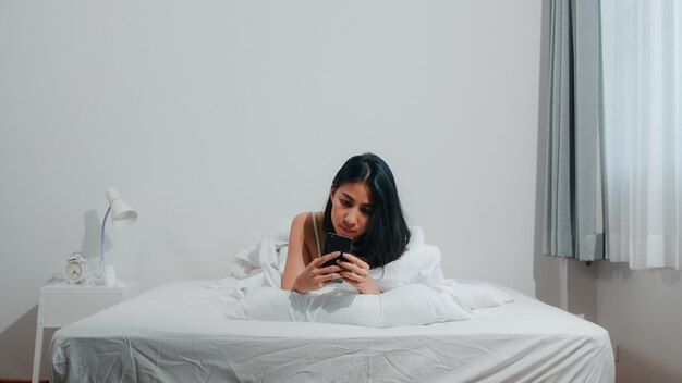 La giovane donna asiatica che utilizza lo smartphone che controlla i media sociali che ritiene sorridere felice mentre si trova sul letto dopo sveglia di mattina, la bella signora ispanica attraente che sorride si rilassa nella camera da letto a casa.