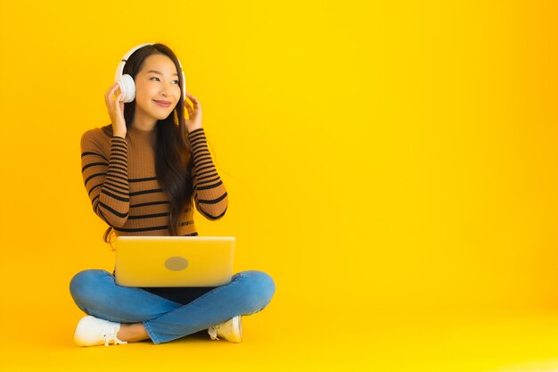 La giovane donna asiatica del bello ritratto si siede sul pavimento con il computer portatile e la cuffia sulla parete gialla
