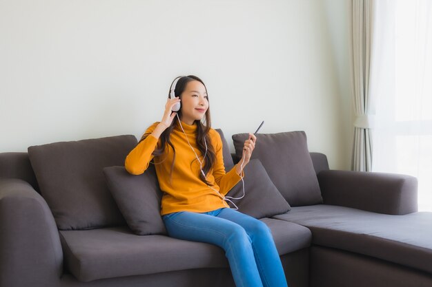 La giovane donna asiatica del ritratto che per mezzo del telefono cellulare astuto con la cuffia per ascolta musica sul sofà